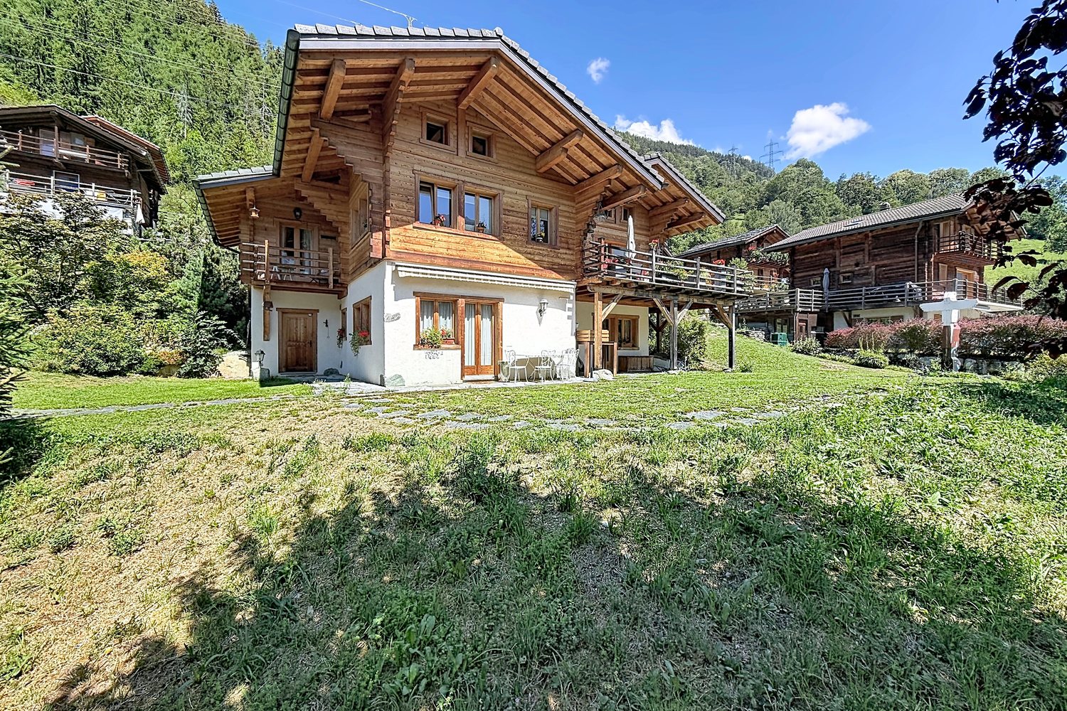 Le chalet vu de l'amont, entouré de pelouse, avec les anciens chalets de bois du hameau en arrière-plan.